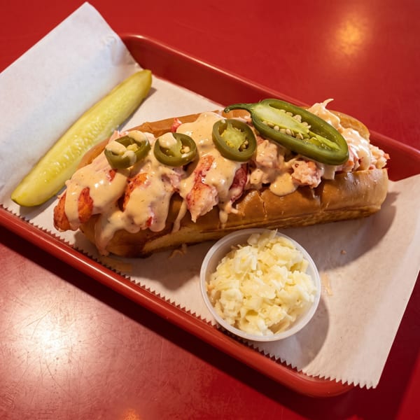 Lobster roll with sauce and jalapeños, served with a pickle spear and coleslaw on a red tray.