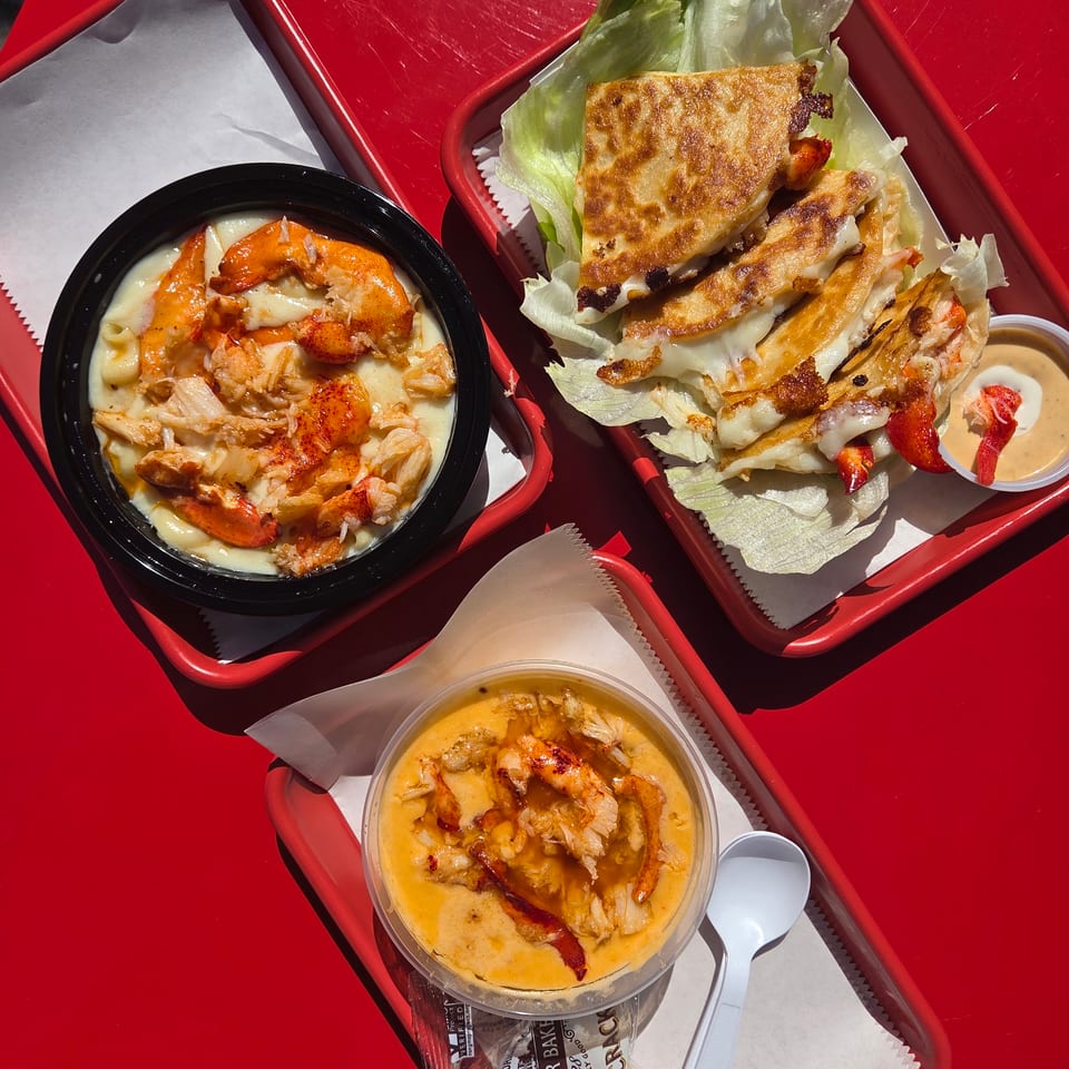 Three lobster dishes on red trays: mac and cheese, quesadillas with lettuce, and a soup with crackers.