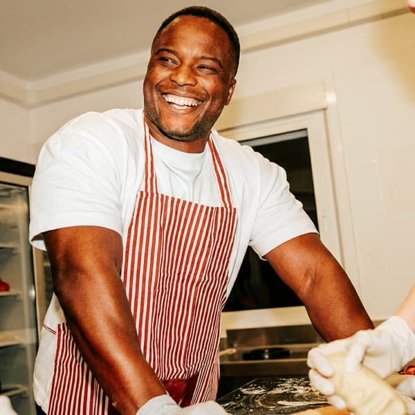 A smiling chef in a red and white striped apron prepares food with others in a kitchen.