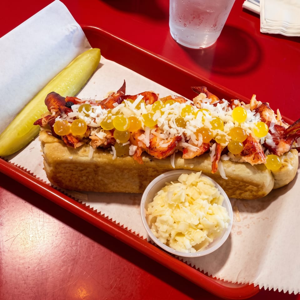 Lobster roll with yellow spheres, shredded white cheese, coleslaw, and a pickle on a red tray.