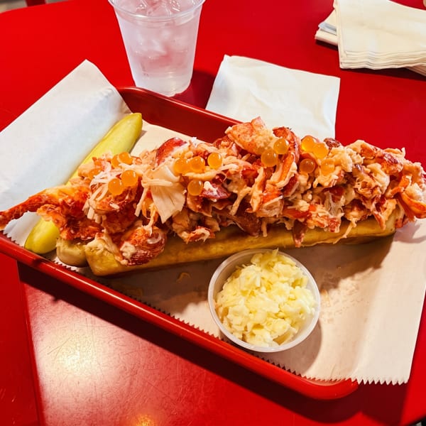 A lobster roll topped with orange roe, served with coleslaw and a pickle on a red tray.
