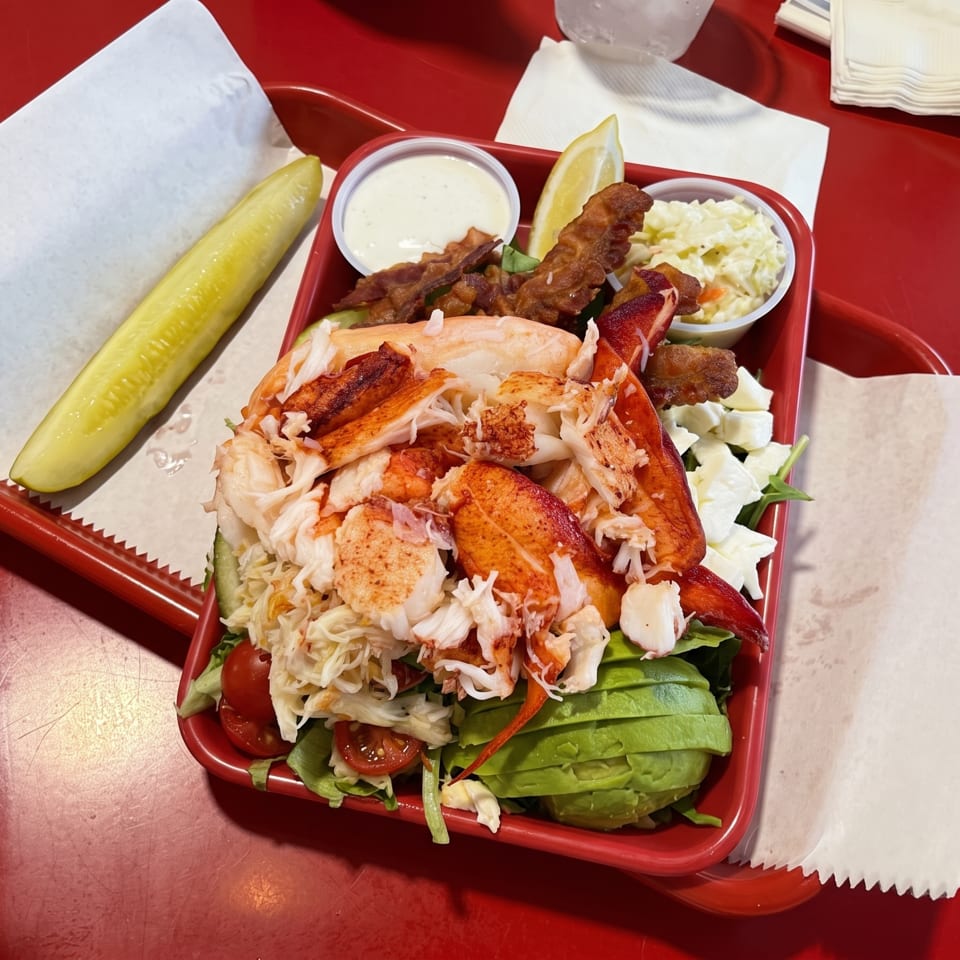 A vibrant lobster salad with avocado, tomatoes, bacon, coleslaw, and a pickle on a red tray.