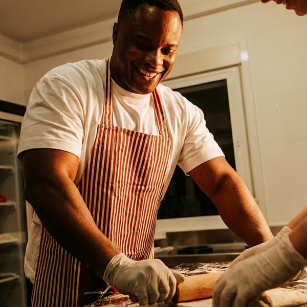 Two chefs in striped aprons and gloves, smiling while rolling dough on a floured surface.