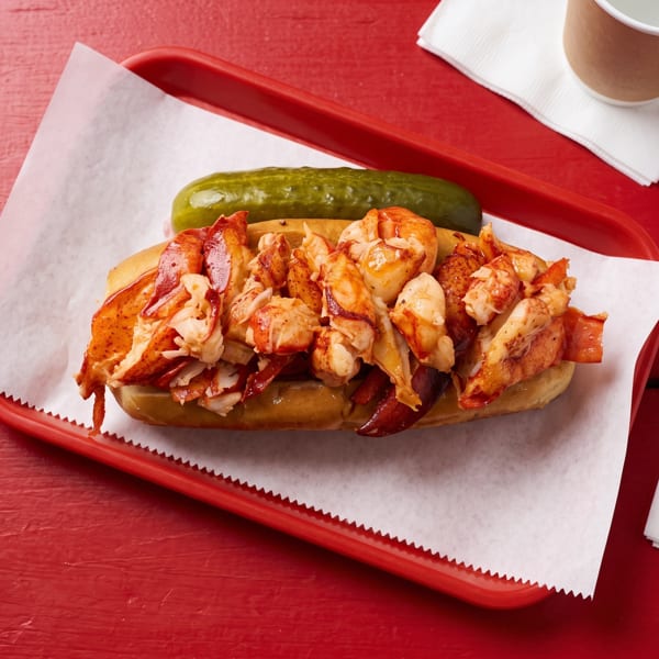 Lobster roll with a pickle spear on a red tray with white paper, napkins, and a drink.