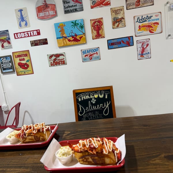 Two lobster rolls with coleslaw on red trays sit on a wooden table, with lobster-themed decor on the wall.