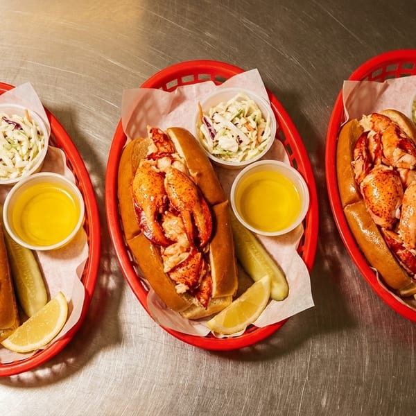 Three lobster rolls in red baskets with coleslaw, butter, pickle, and lemon on a metal table.