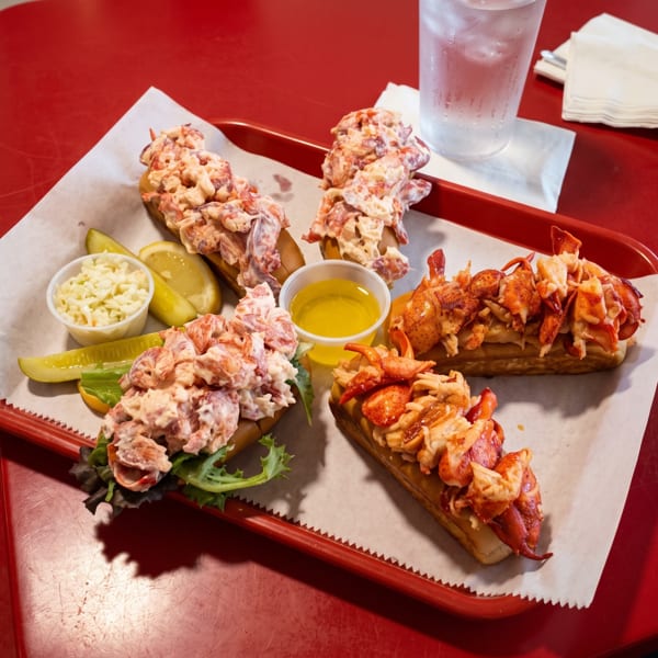 Four lobster rolls, coleslaw, pickles, lemon, and butter on a red tray with a drink.
